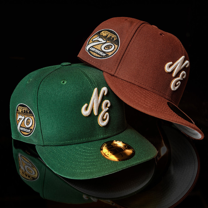 two fitted 59FIFTY caps, one green and one brown, with NE scripted on the front and a 70 years anniversary badge on the right